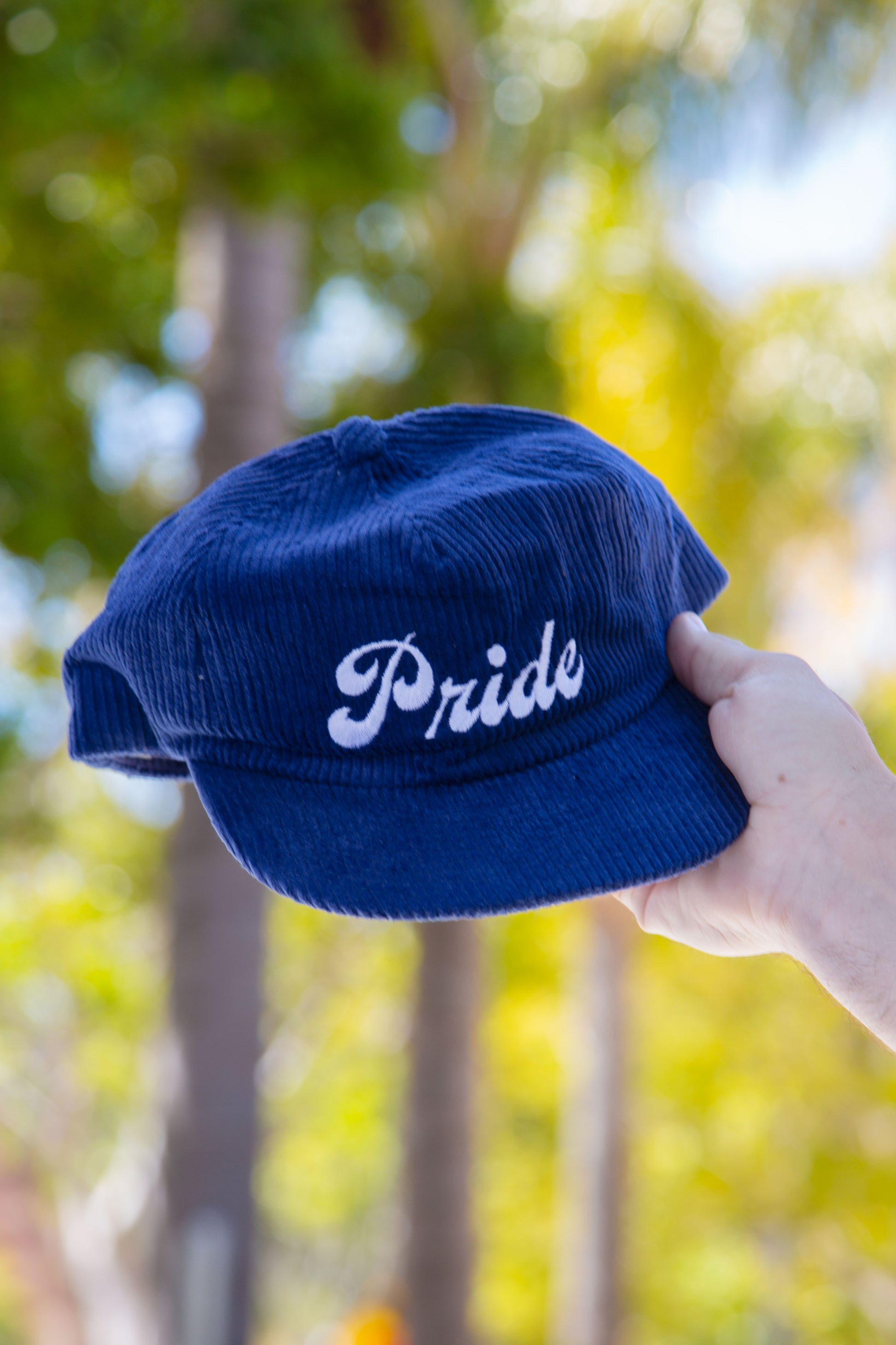 Person holding the navy blue <Insert Material Type> hat with the text "Pride" on the front outside with a blurred background.
