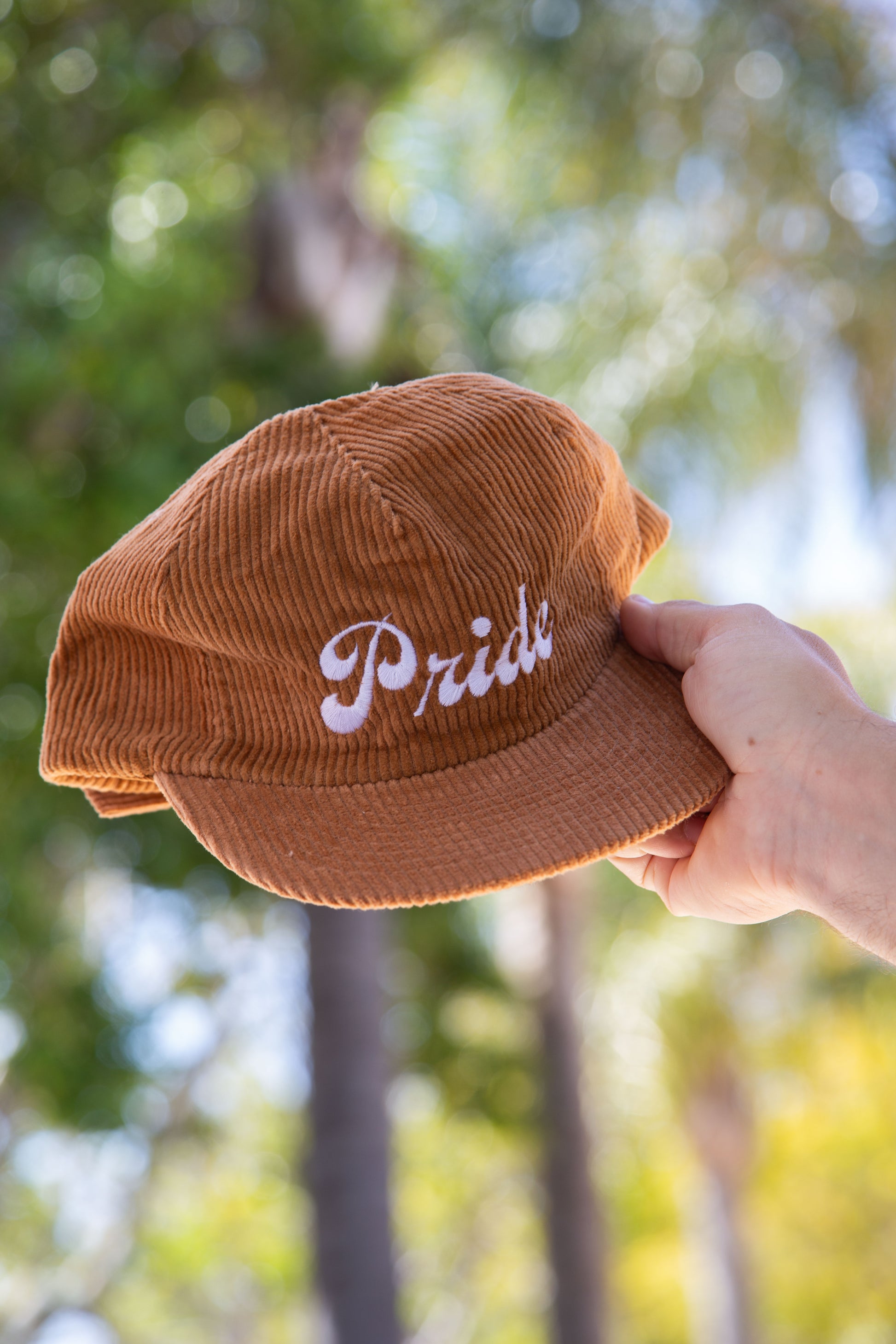Person holding the brown <Insert Material Type> hat with the text "Pride" on the front outside with a blurred background.