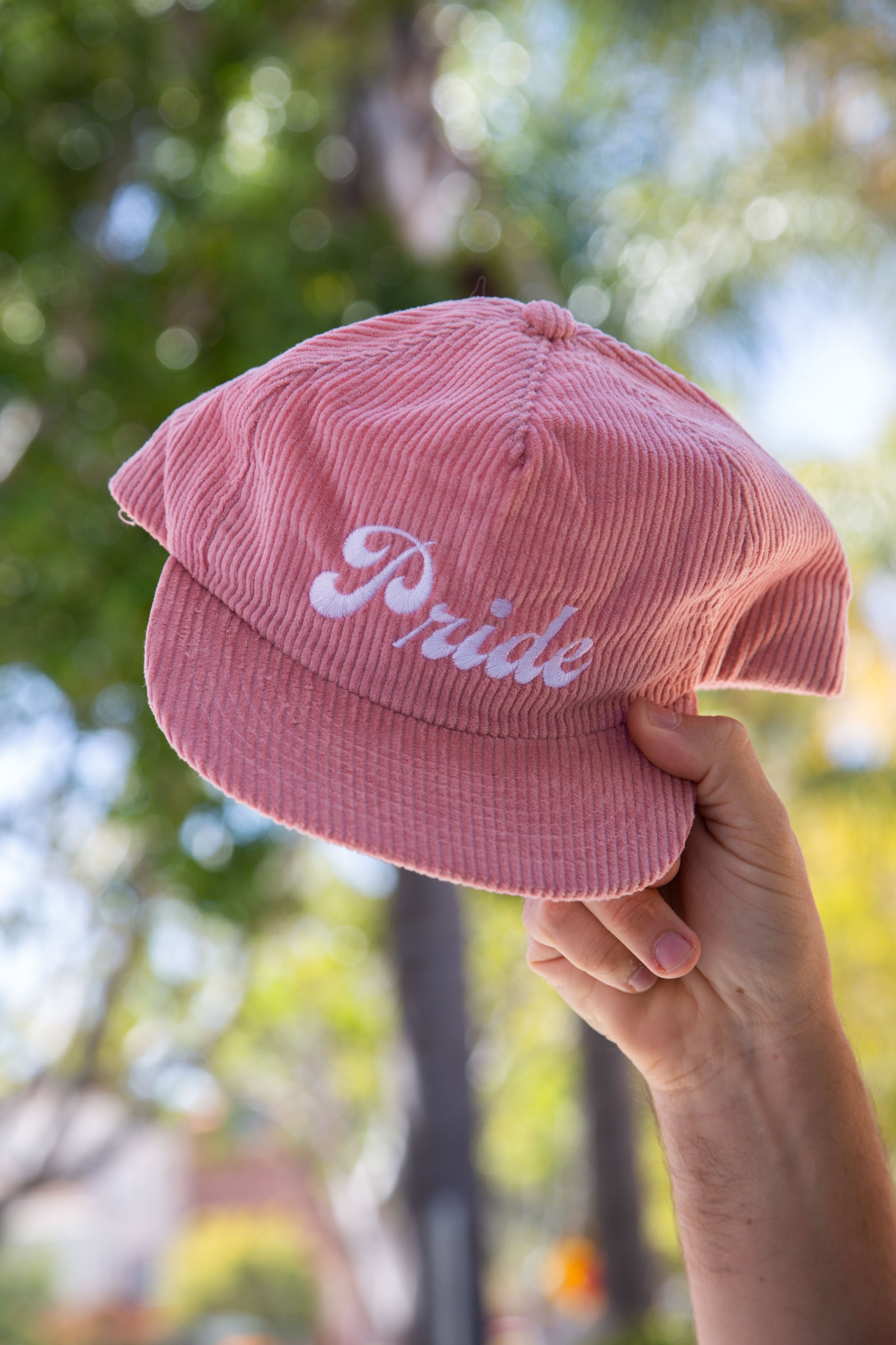 Person holding the pink <Insert Material Type> hat with the text "Pride" on the front outside with a blurred background.